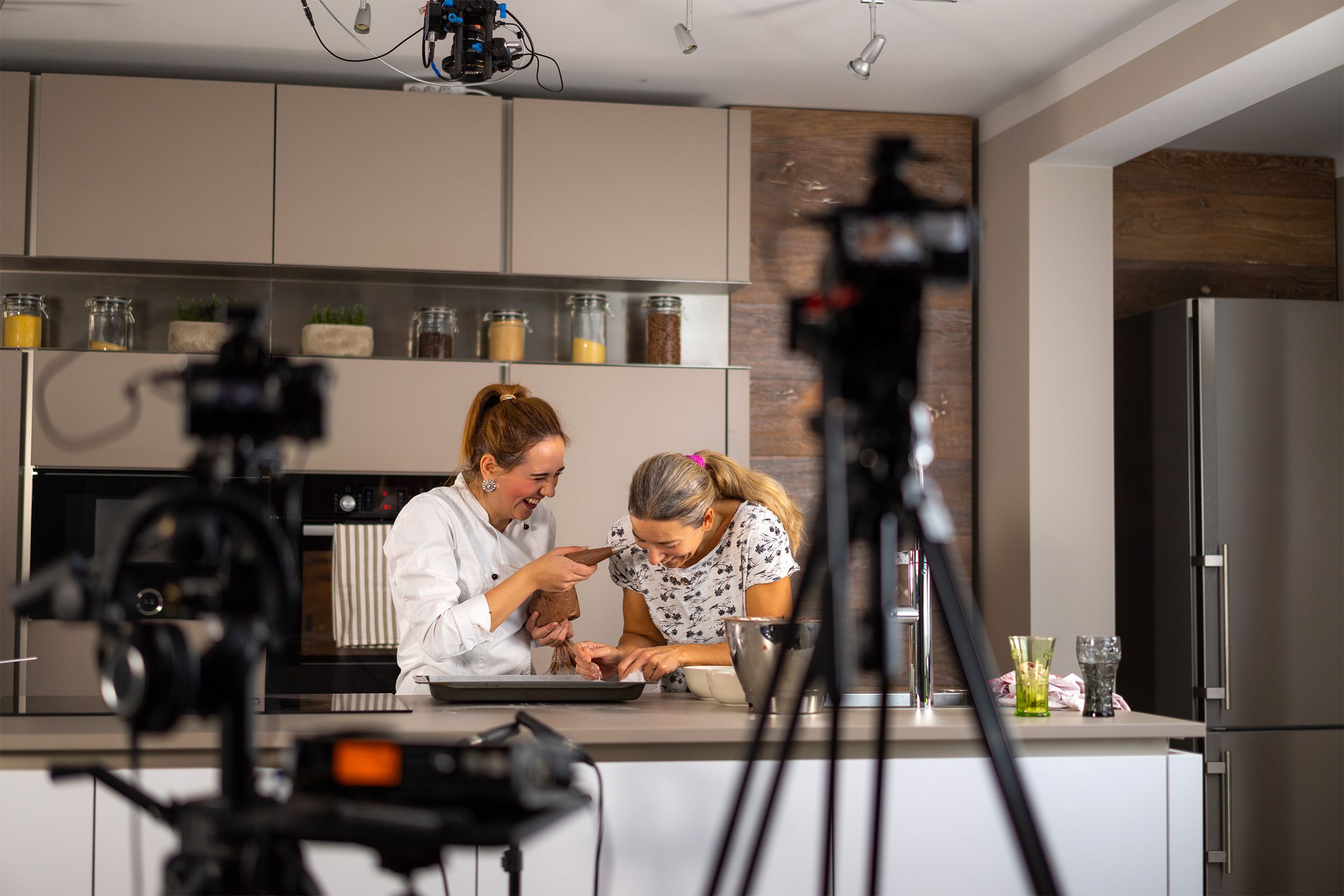 Vlog movie equipment blurred in the foreground with two senior woman having fun cooking in a home kitchen in the background