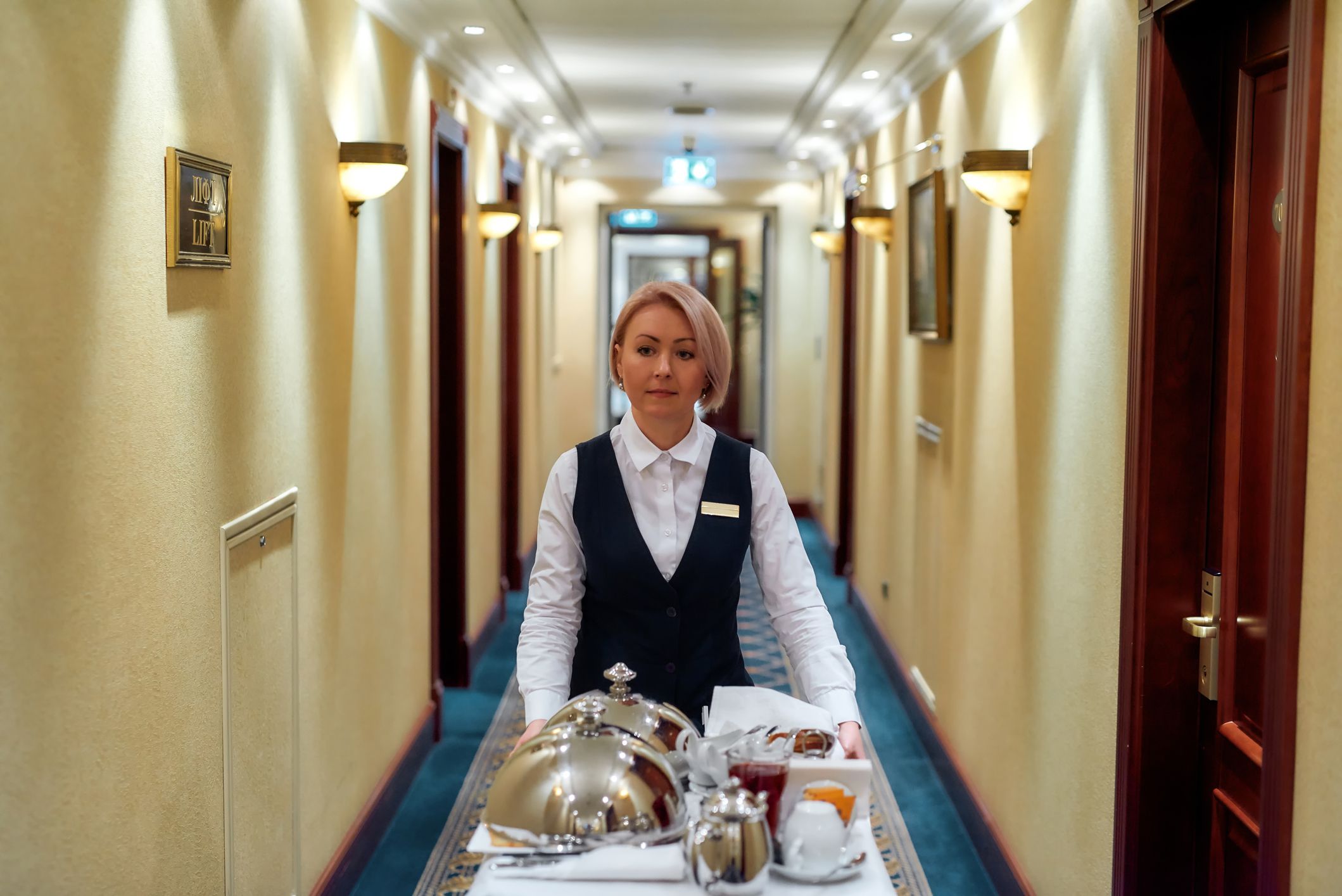 Woman working in an upscale hotel, dressed in a uniform, walking down the hall of rooms delivering a tray with food for room service, end of the hallway blurred in the background