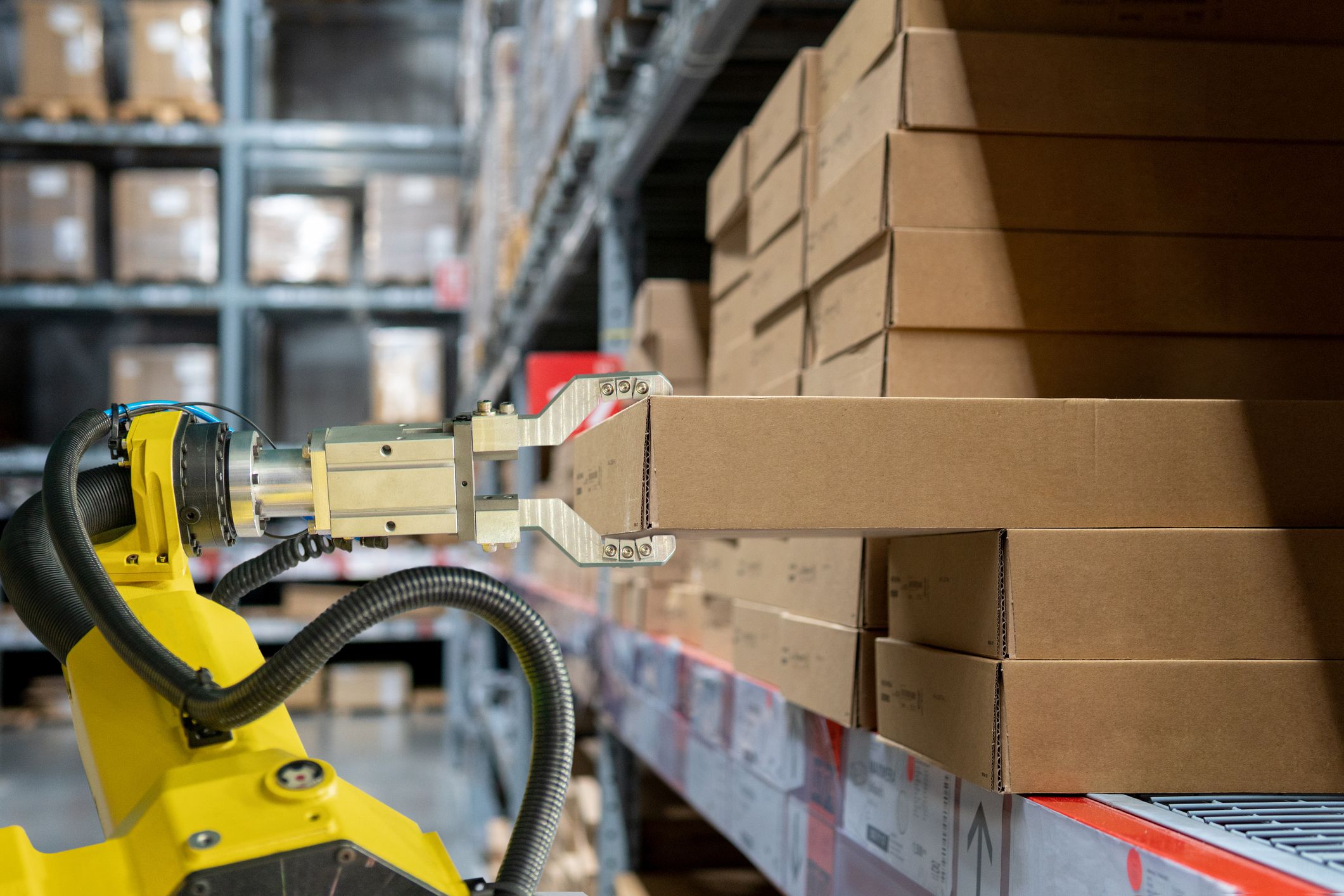 Closeup of yellow robotic arm is pulling out a cardboard box in warehouse from stack of cardboard boxes, other rows of cardboard boxes in the background, blurred