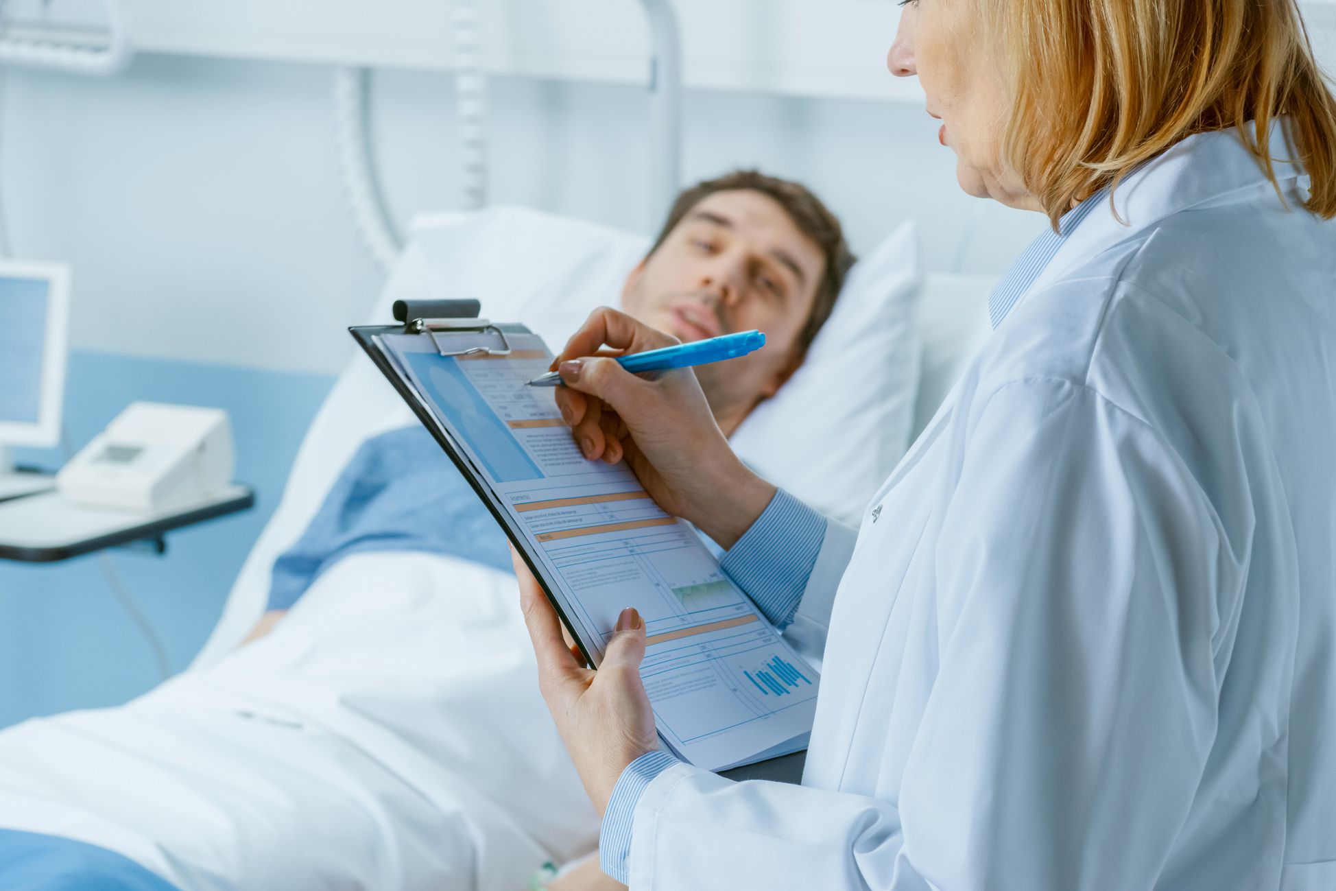 man in hospital bed speaking to doctor who reviews chart