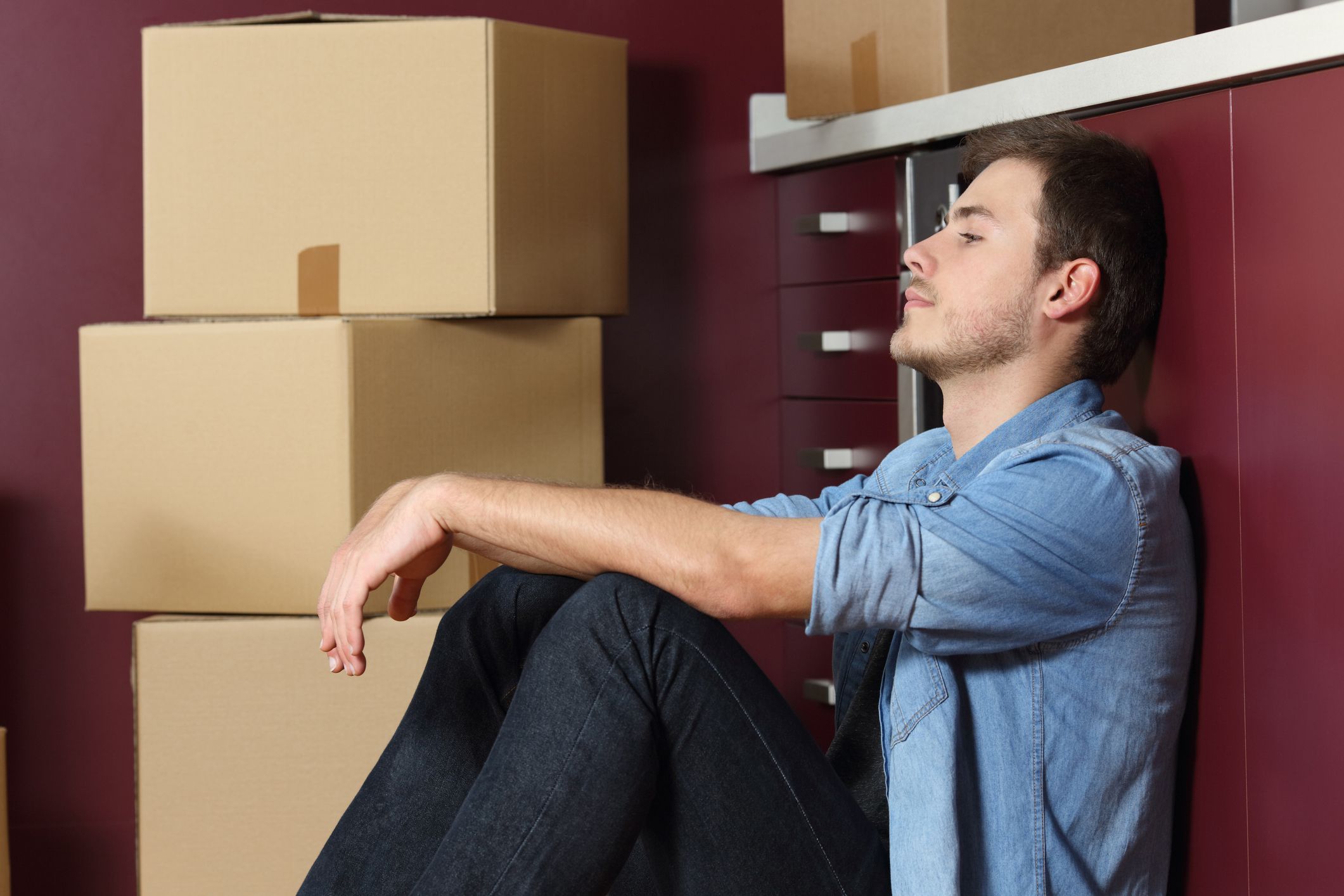 sad man sitting on floor leaning against counter, cardboard boxes stacked next to him