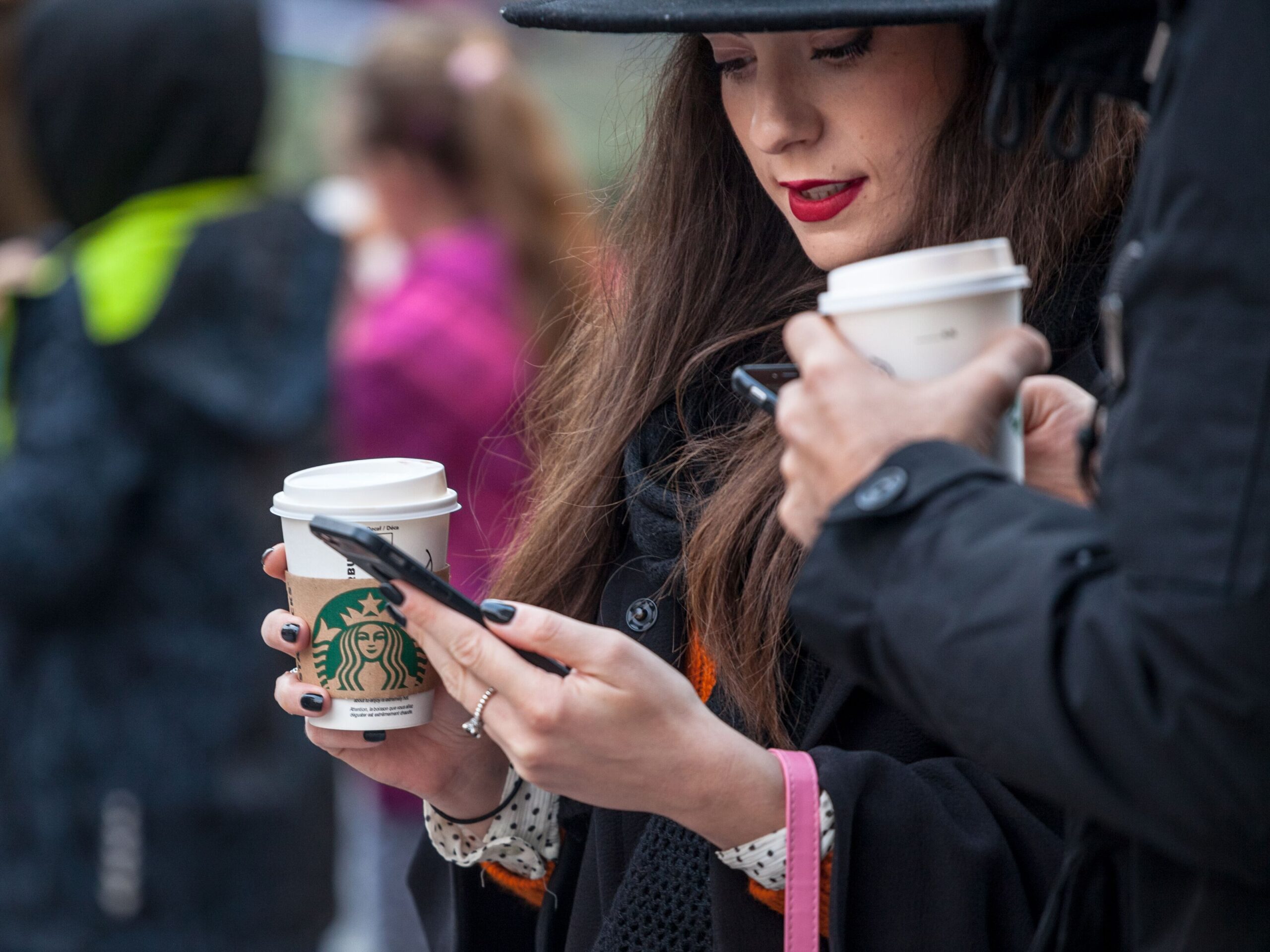 Picture of a young woman holding her smartphone while drinking a coffee in a cardboard coffee cup with the logo of Starbucks. Starbucks Corporation is an American coffee company and coffeehouse chain operating worldwide