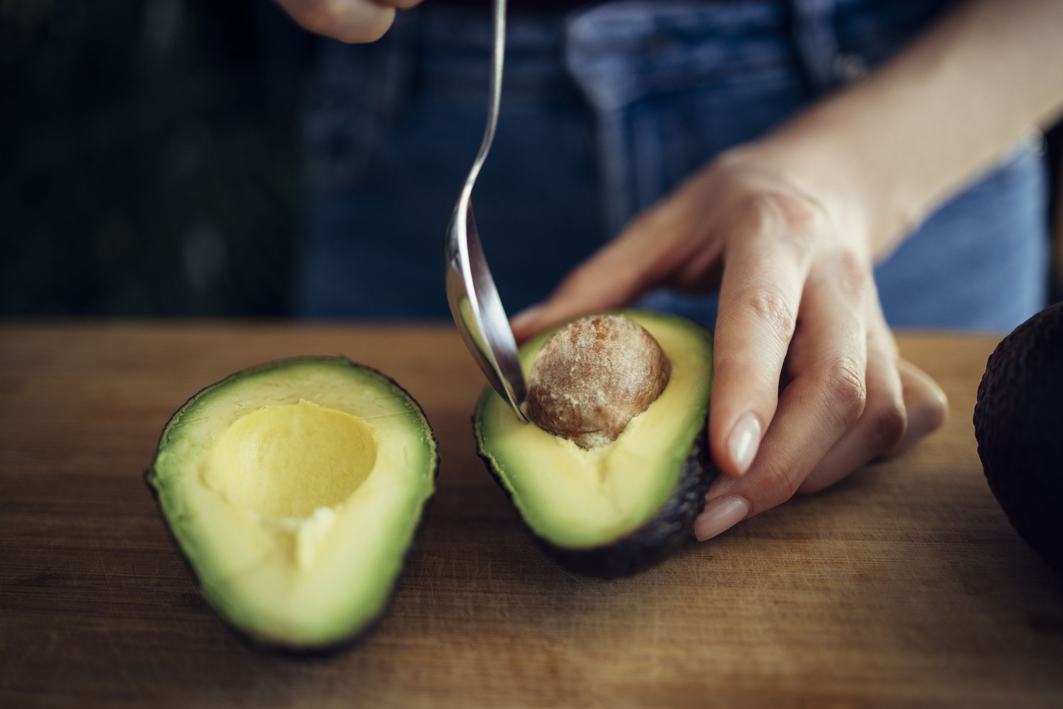 Unrecognizable female person peeling perfectly ripe avocado with spoon on wooden cutting board in kitchen
