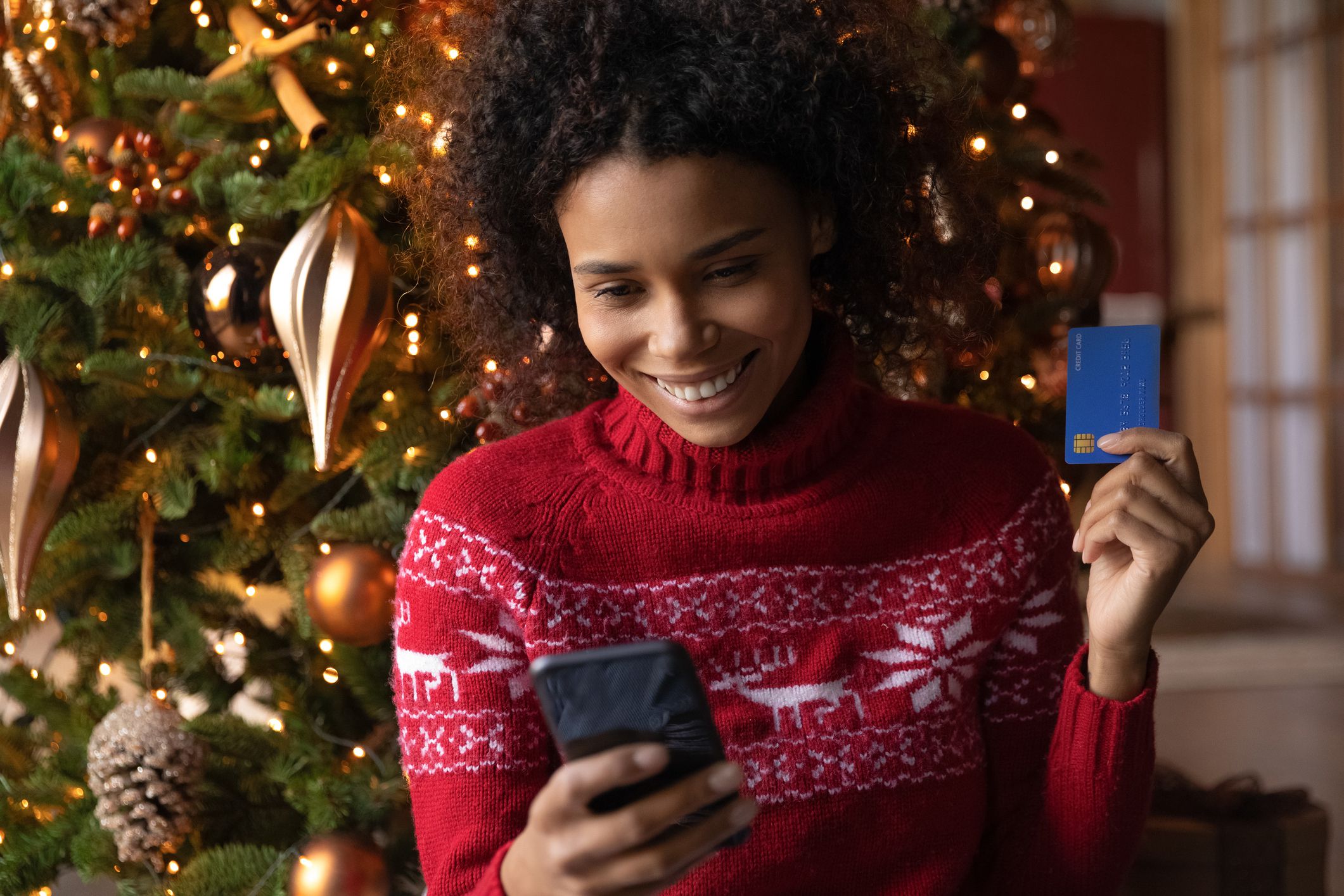 Happy young African American woman shop online for Christmas presents with credit debit card on smartphone. Smiling biracial female buy New Year gifts on winter holiday sale on internet on cellphone.