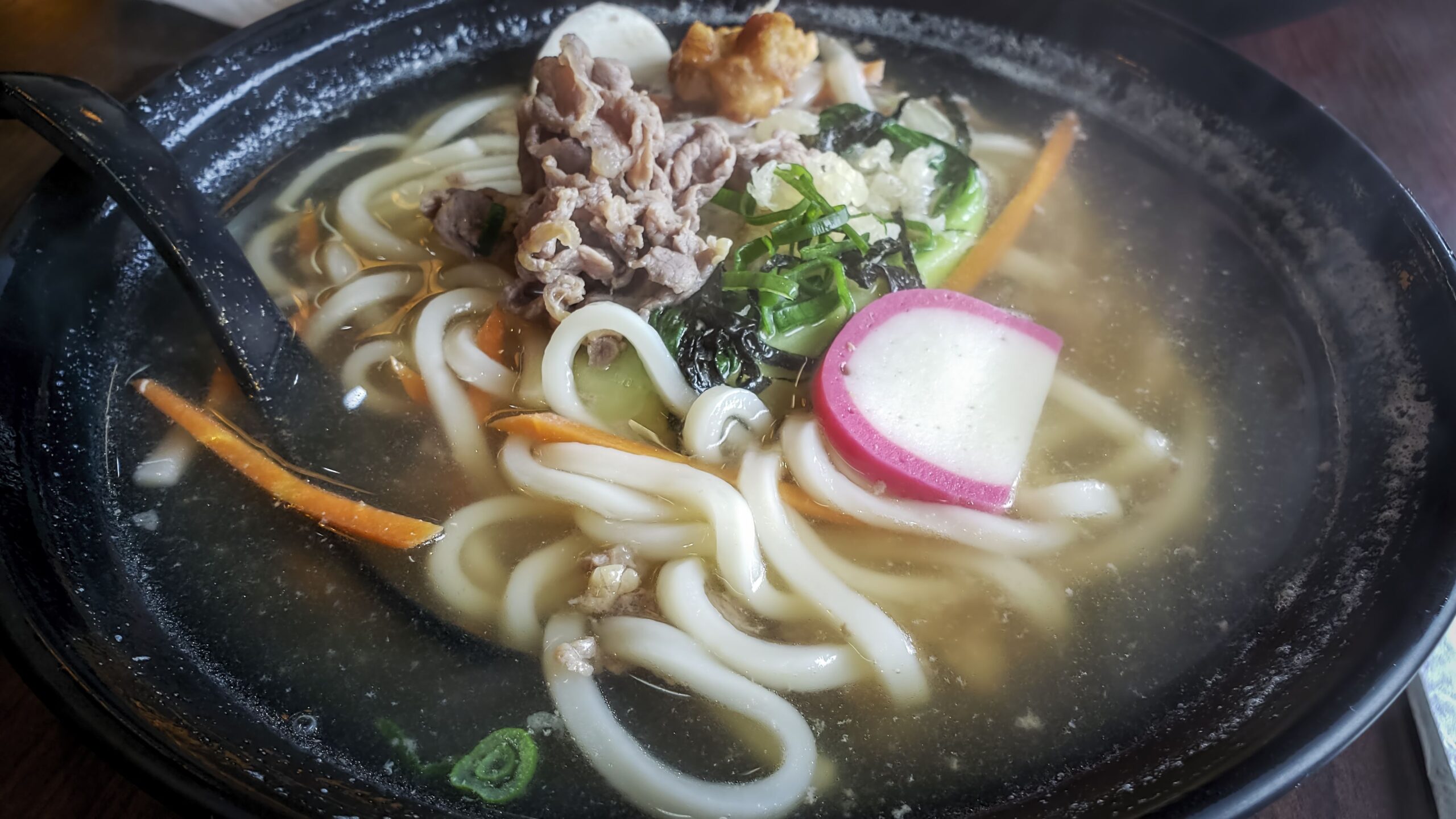 Healthy bowl of udon noodles in hot soup, with thinly sliced beef pieces, and garnished with vegetables and seafood. Rising hazy steam. Popular Japanese cuisine. A catering concept.