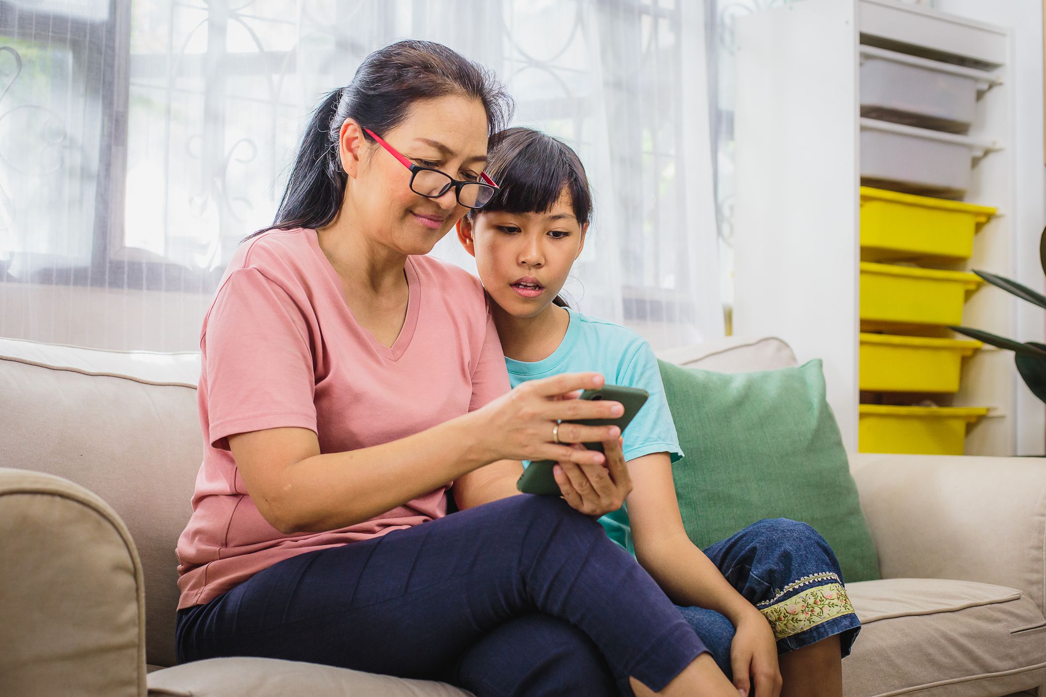 Happy Asian mother and her teenage daughter using smart phone while sitting on sofa in living room at home, Family relaxing and spending time together at home