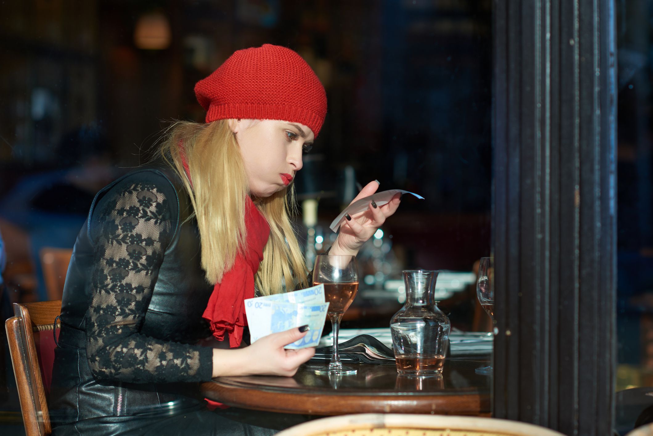 side view of young woman with wine glass on the table reading the check note and having displeased expression.