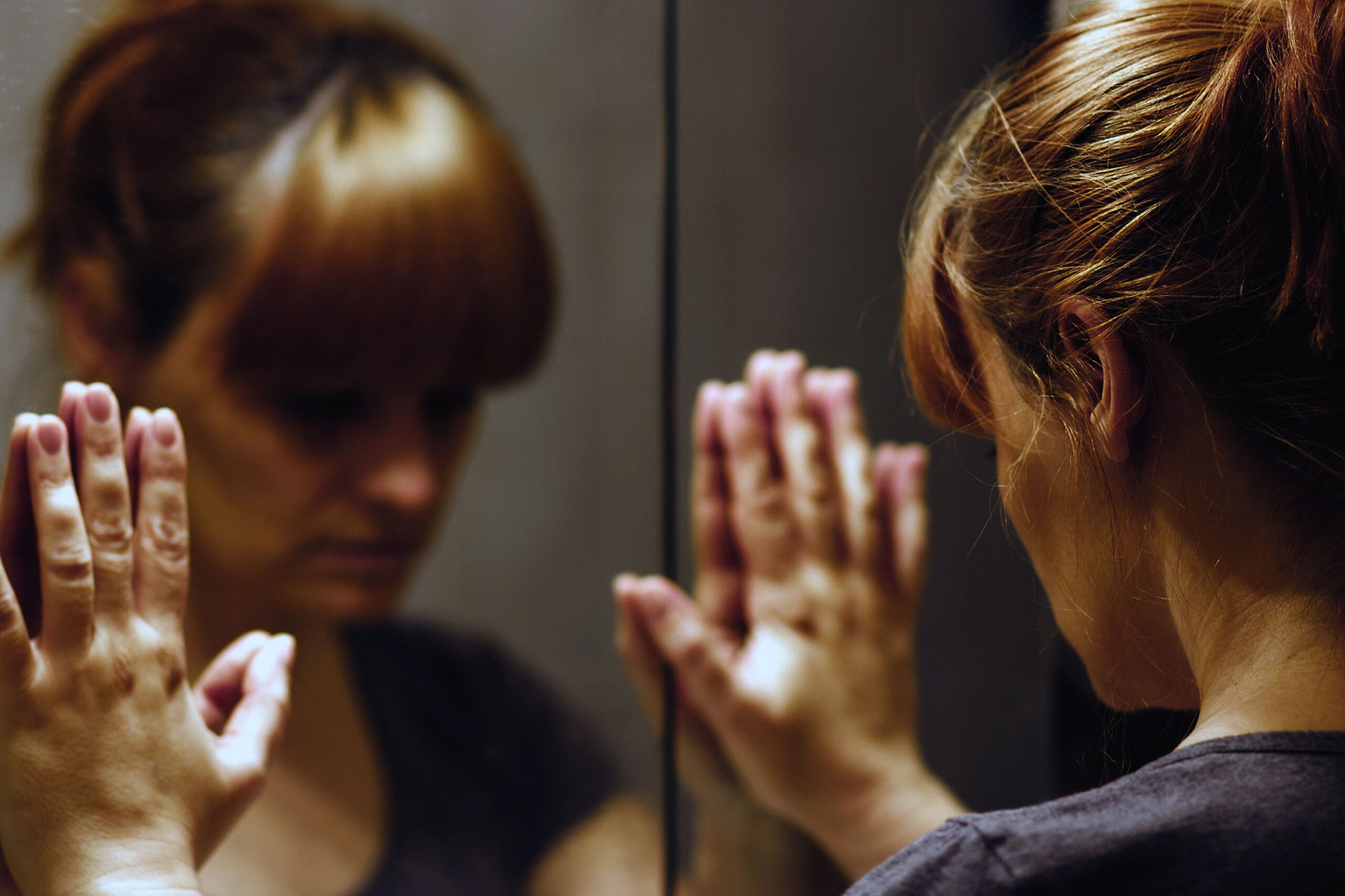 woman looking at reflection in mirror, distressed, with hands placed against mirror