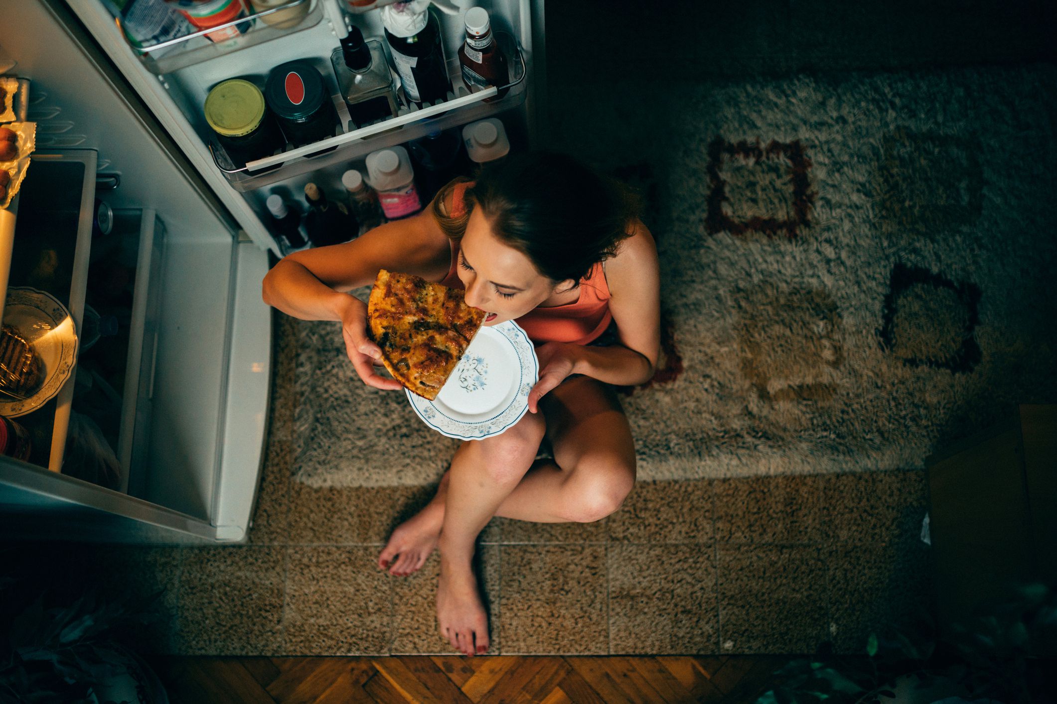 woman sitting on floor with open fridge eating pizza