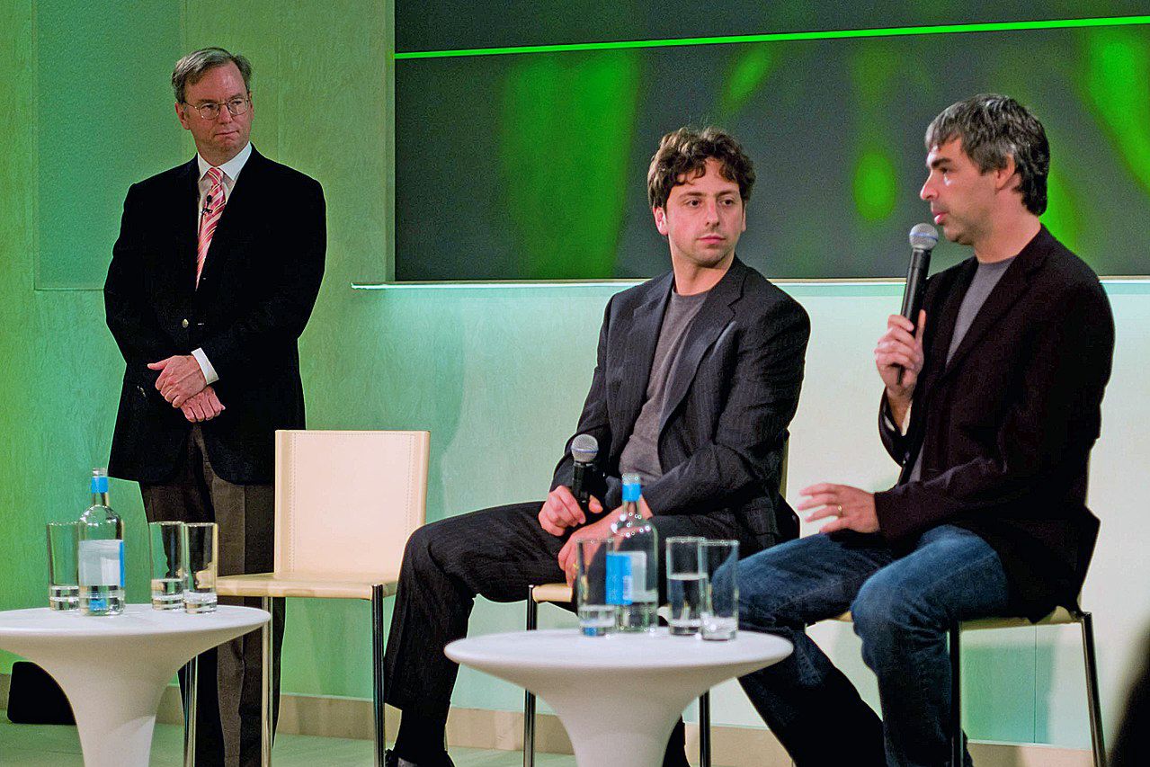 Three men are at an event; two are seated on stools with microphones, talking, while one stands to the side watching. Water bottles and glasses are on the tables. The setting is modern with green lighting in the background.
