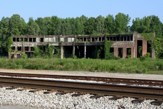 An abandoned brick building with broken windows and overgrown vegetation stands behind a set of train tracks and gravel, with trees in the background under a clear sky.