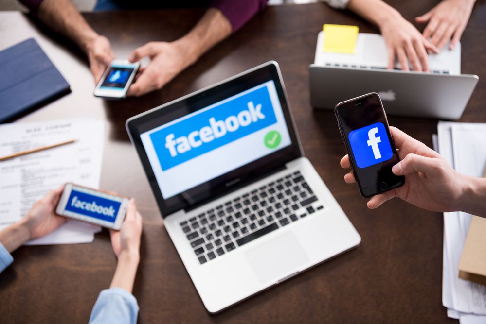 Four people sitting at a table use various devices, including a laptop, smartphones, and a tablet, all displaying the Facebook logo or website.