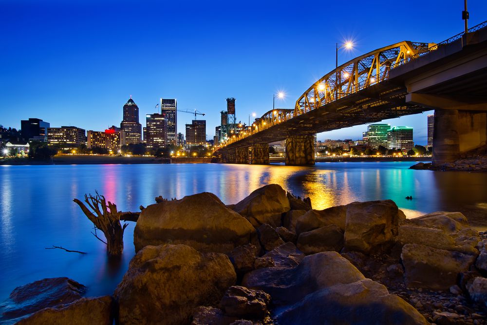 A city skyline at dusk with bright lights reflecting on the water, a lit bridge spanning the river, and large rocks in the foreground under a deep blue sky.