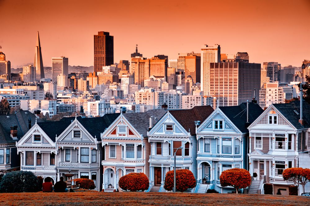 A row of colorful Victorian houses, known as the Painted Ladies, stands in front of the San Francisco skyline at sunset, with warm orange light casting over the buildings and sky.