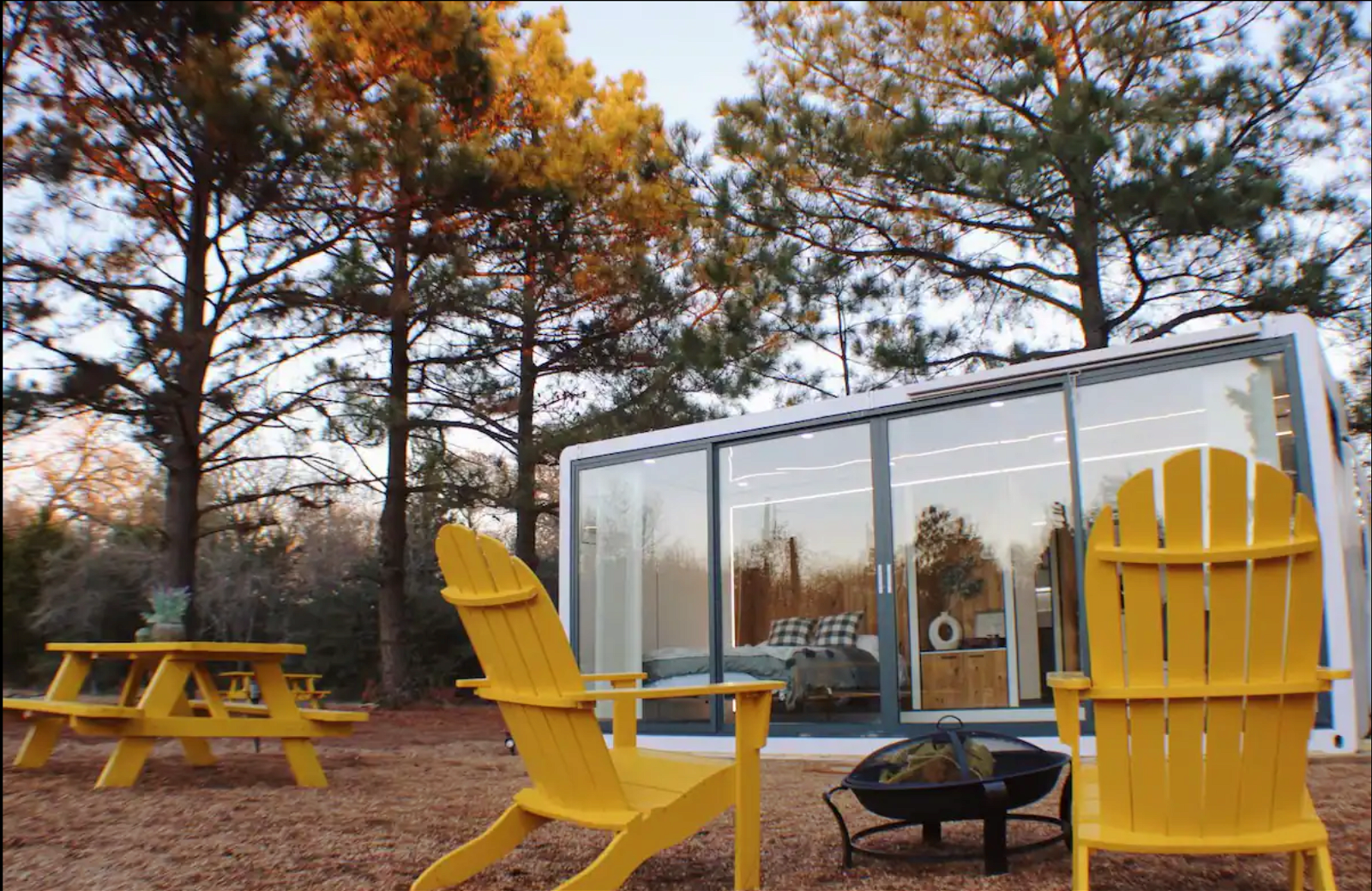 Front exterior of Expandable Tiny House, Plano, Texas, large windows, two yellow chairs and matching yellow park table in the foreground, on mulch and grass surrounded by large trees during late autumn