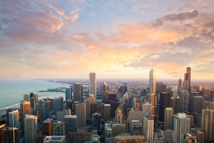 Aerial view of a city skyline with tall skyscrapers at sunset, with a large body of water on the left and a colorful sky with clouds above.