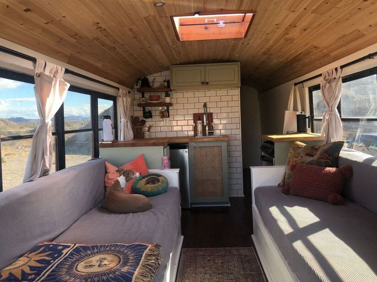 Interior of Gorgeous Off-Grid Skoolie, North Port, Florida, two couches on both sides, wooden ceiling, and a kitchen nook in the background, on a sunny day with mountains through the windows