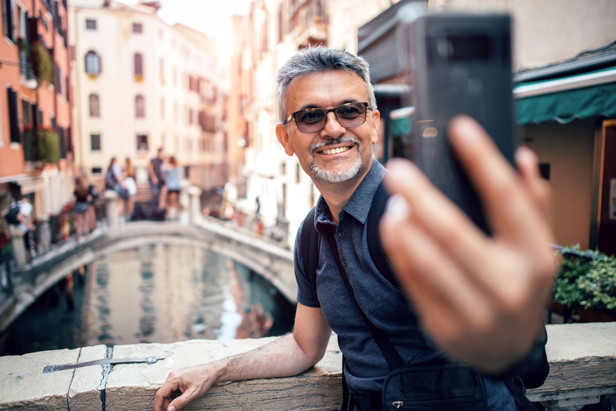 Happy senior man smiling while taking selfie picture with his smartphone in Venice, Italy, he is resting his arm on a ledge with a river in the background