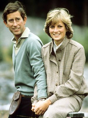 A man and woman sit close together outdoors, holding hands and smiling. The man wears a collared shirt and kilt, while the woman wears a checked jacket and trousers. The background is blurred greenery.