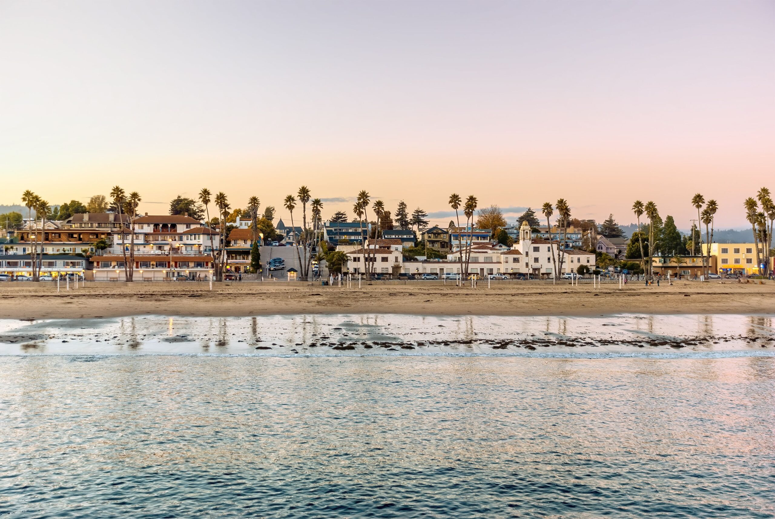 Focus on ocean in Santa Cruz, California during sunset with beach, houses, and palm trees in the background