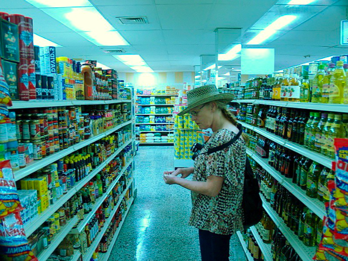 A woman wearing a hat and patterned shirt stands in a grocery store aisle, looking at a small item in her hands, surrounded by shelves stocked with various food products.