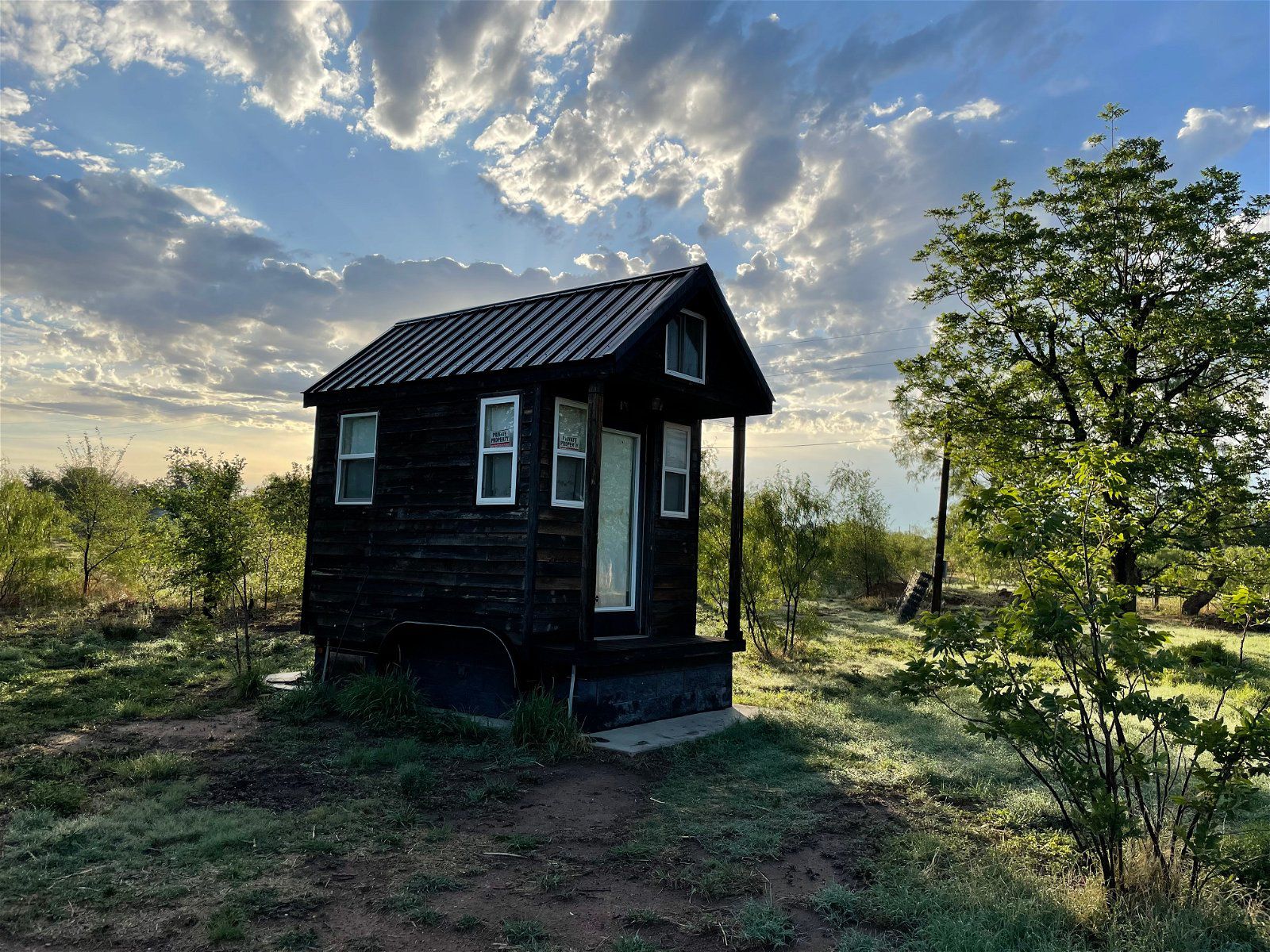 Front and side exterior of Tiny Home With 1.2 Acres, Spur, Texas, on a grassy lot with bushes and trees, against a dramatic sunny sky during summer