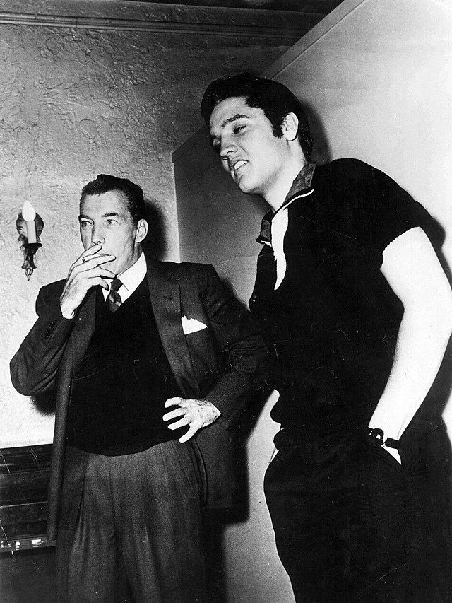 Two men stand indoors; one in a suit smokes a cigarette with a thoughtful expression, while the other, dressed casually in dark clothing, stands beside him with hands in his pockets, both looking in the same direction.