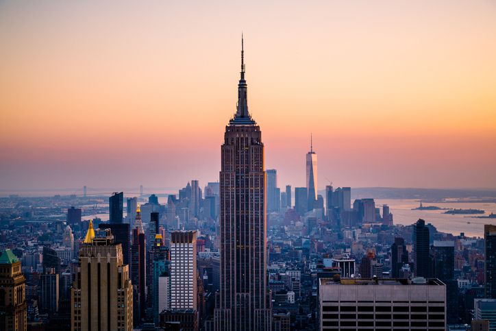 The Empire State Building stands tall in the foreground with the New York City skyline and distant skyscrapers, all under a colorful sunrise or sunset sky.
