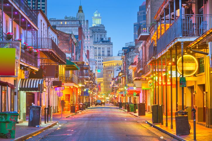 A vibrant street in New Orleans' French Quarter at dusk, lined with colorful buildings, neon signs, balconies, and glowing lights, with a city skyline in the background.