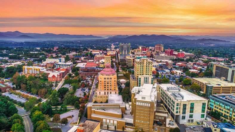 Aerial view of a city with colorful buildings surrounded by greenery, set against a backdrop of rolling mountains under an orange and pink sunset sky.