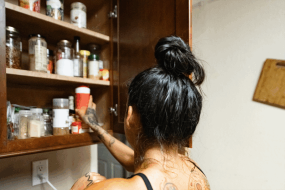 A person with their hair in a bun reaches into an open kitchen cupboard to retrieve a container.