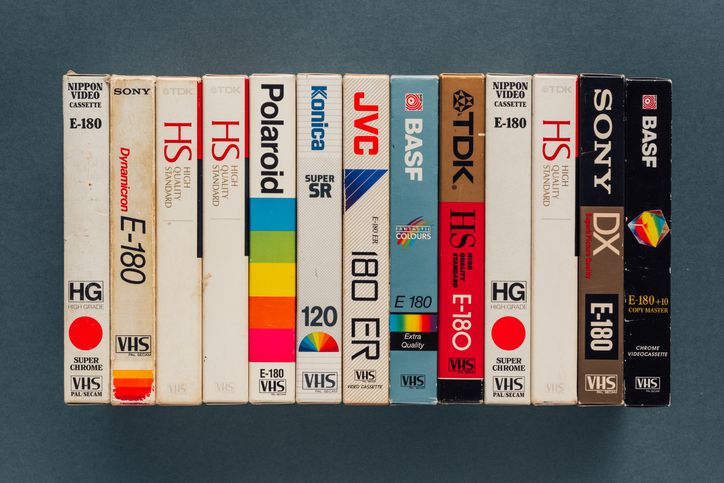 A row of twelve vintage VHS tapes, each with different branded labels and designs, standing upright side by side against a dark background.