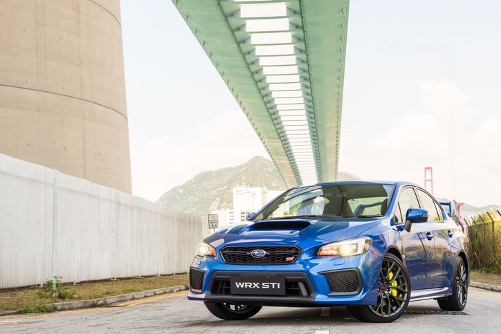 A blue Subaru WRX STI car is parked on a street under a large modern bridge, with mountains and a partially visible suspension bridge in the background.
