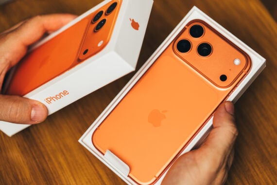 A person holds an orange iPhone with three cameras in its box, next to the phone's packaging, on a wooden surface—highlighting common Black Friday shopping mistakes like rushing expensive tech buys.