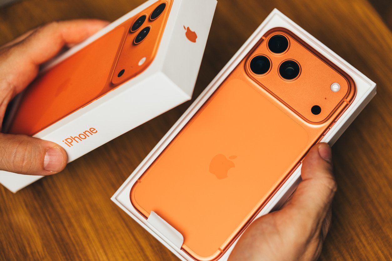 A person holds an orange iPhone with three cameras in its box, next to the phone's packaging, on a wooden surface—highlighting common Black Friday shopping mistakes like rushing expensive tech buys.