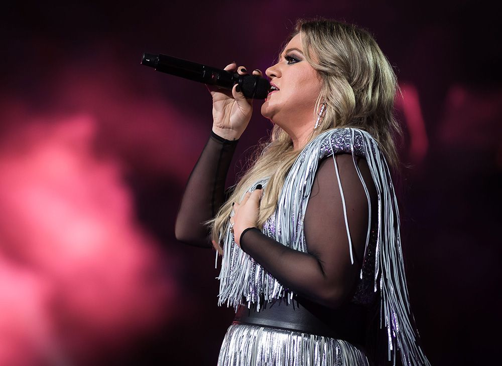 A woman with long blonde hair sings into a microphone on stage, wearing a sparkly, fringed outfit with sheer sleeves, against a pink and purple smoky background.