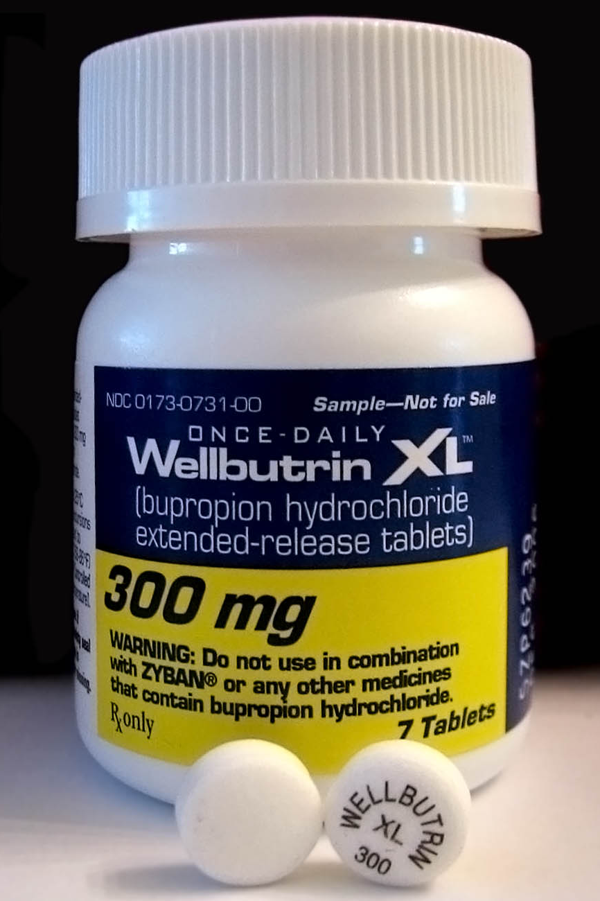 Closeup of an official white pharmacy sample bottle of 300mg Wellbutrin, 7 tablets, two white round tablets in front of bottle on a white table with a black background