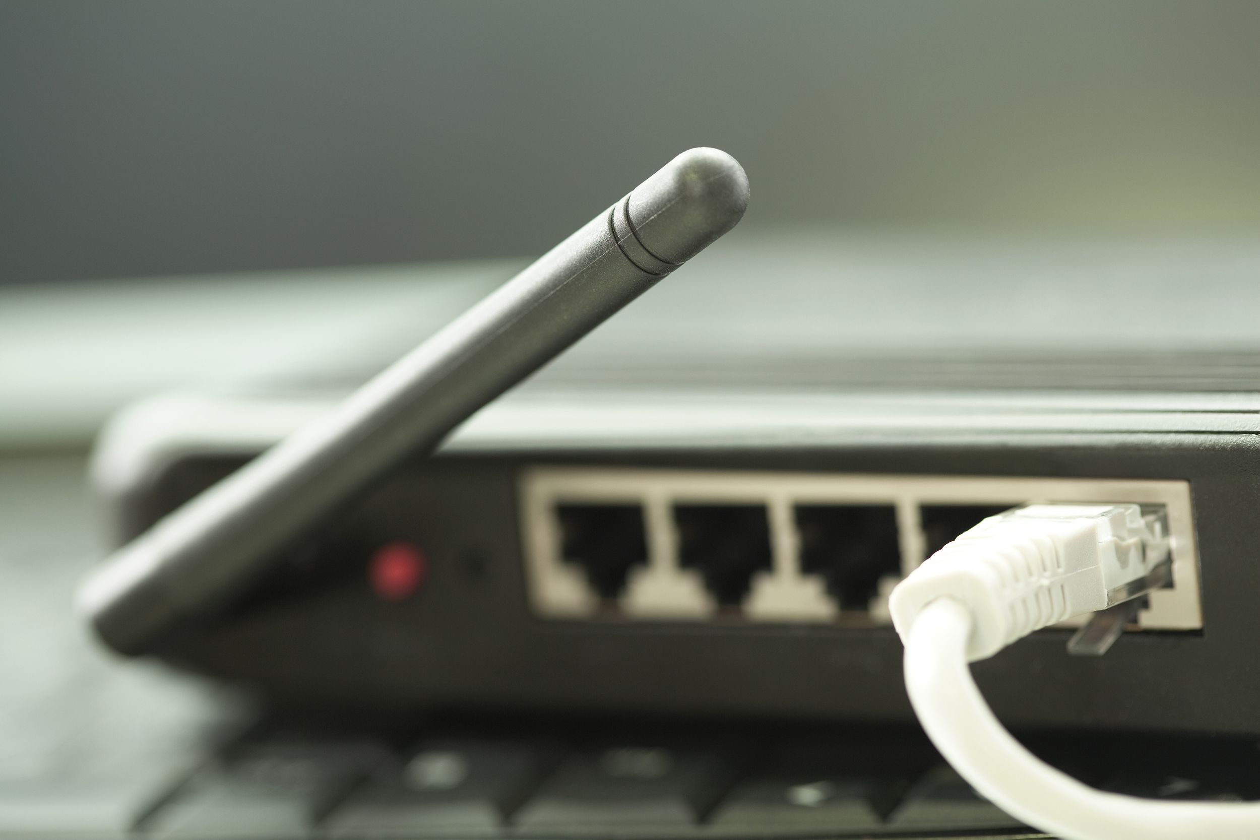 Closeup of back of internet router, antenna and white cable in device, both are selective focus, red light is one, blurred on keyboard and surrounded by grey blur