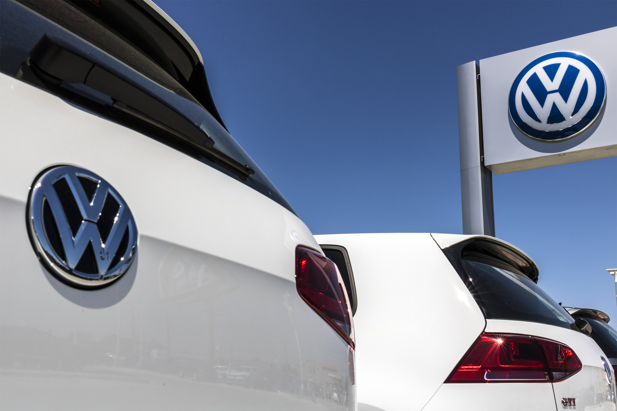 Closeup of back of a white Volkswagen SUV on the left with another white Volkswagen SUV on the left, slight side-view, Volkswagen dealership car sign on the top right, against a vivid blue sky
