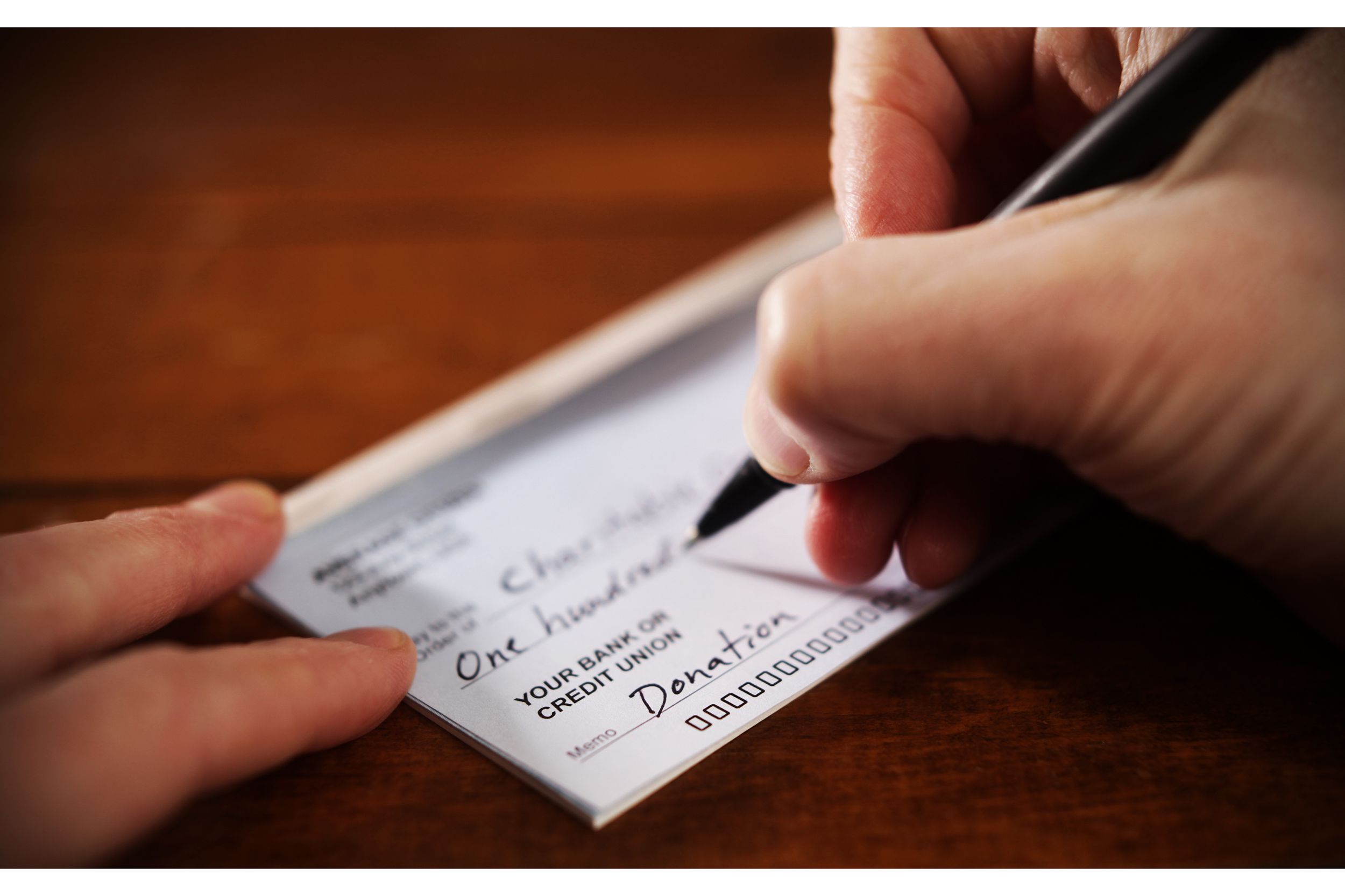 Closeup of a hand holding a pen filling out a check for a donation, selective focus, on a dark wooden table, blurred