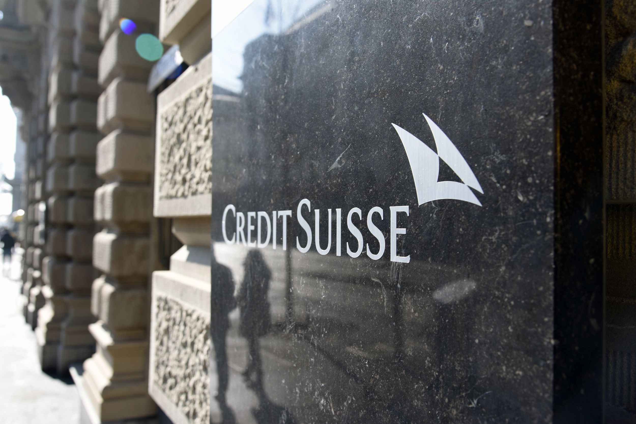 Closeup of sign on black stone on front exterior of headquarters of the Credit Suisse, Zurich, sign in the foreground on the right, beige stone building columns and street blurred in the background on the left