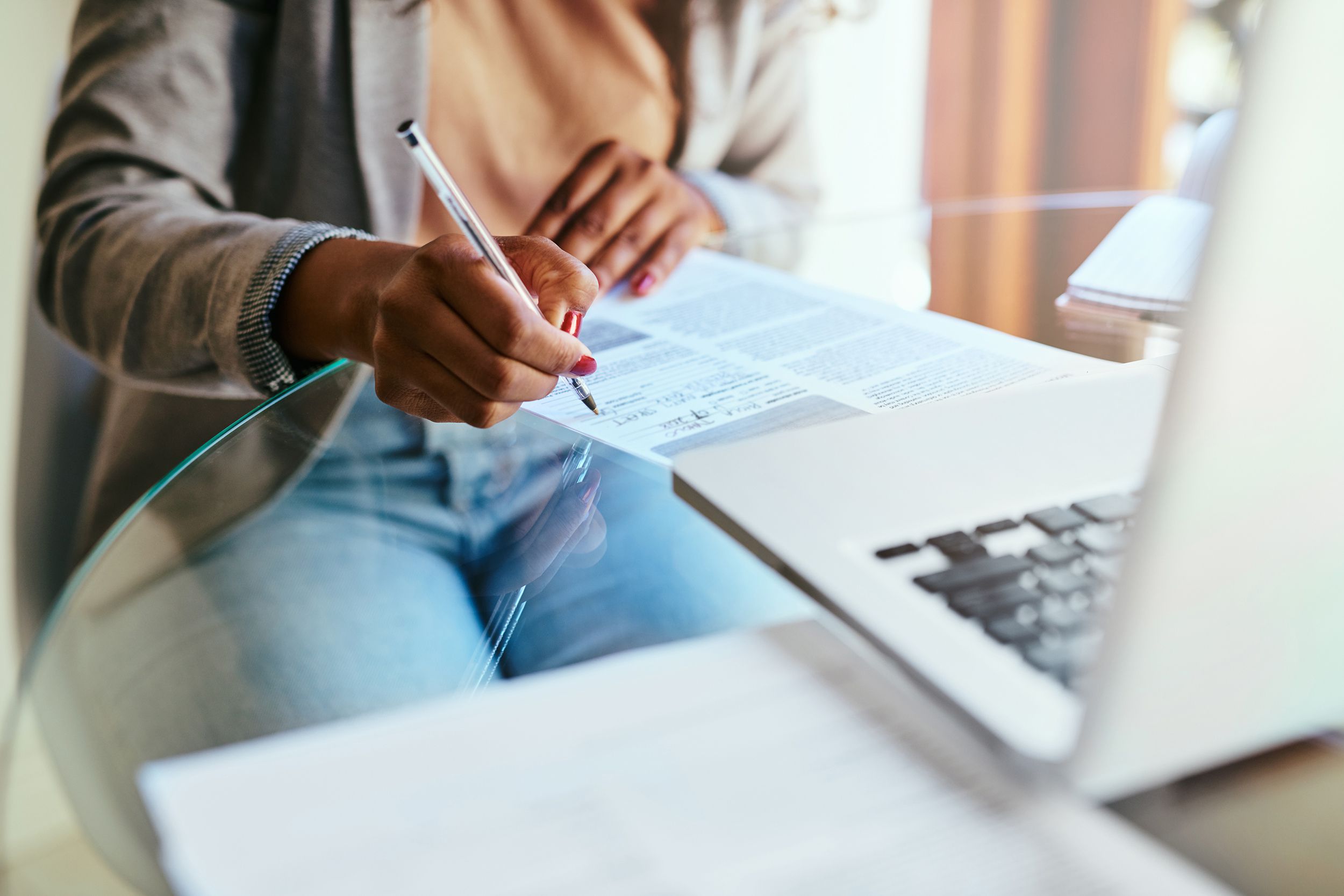 Cropped shot of a woman filling out paperwork on a glass tabletop at home, selective focus, MacBook in the foreground on the right with papers on the left, blurred foreground and background