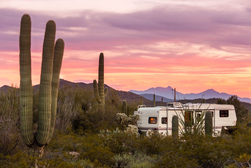A camper trailer is parked among tall saguaro cacti in a desert landscape at sunset, with colorful skies and distant mountains in the background.