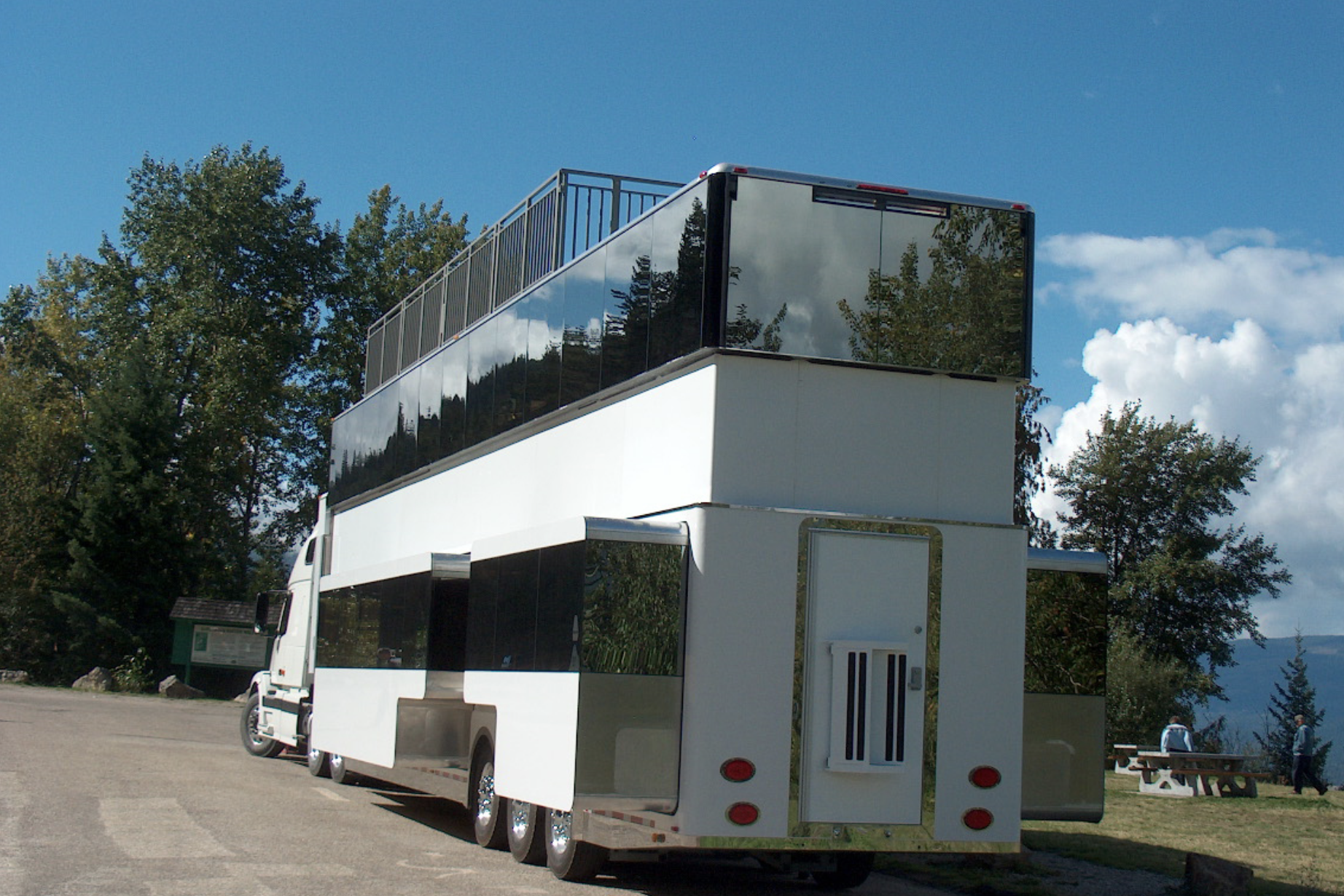 Back and side exterior of Anderson Mobile Estates Anderson Collection RV on a paved parking area in a park, surrounded by a park with mountains in the background