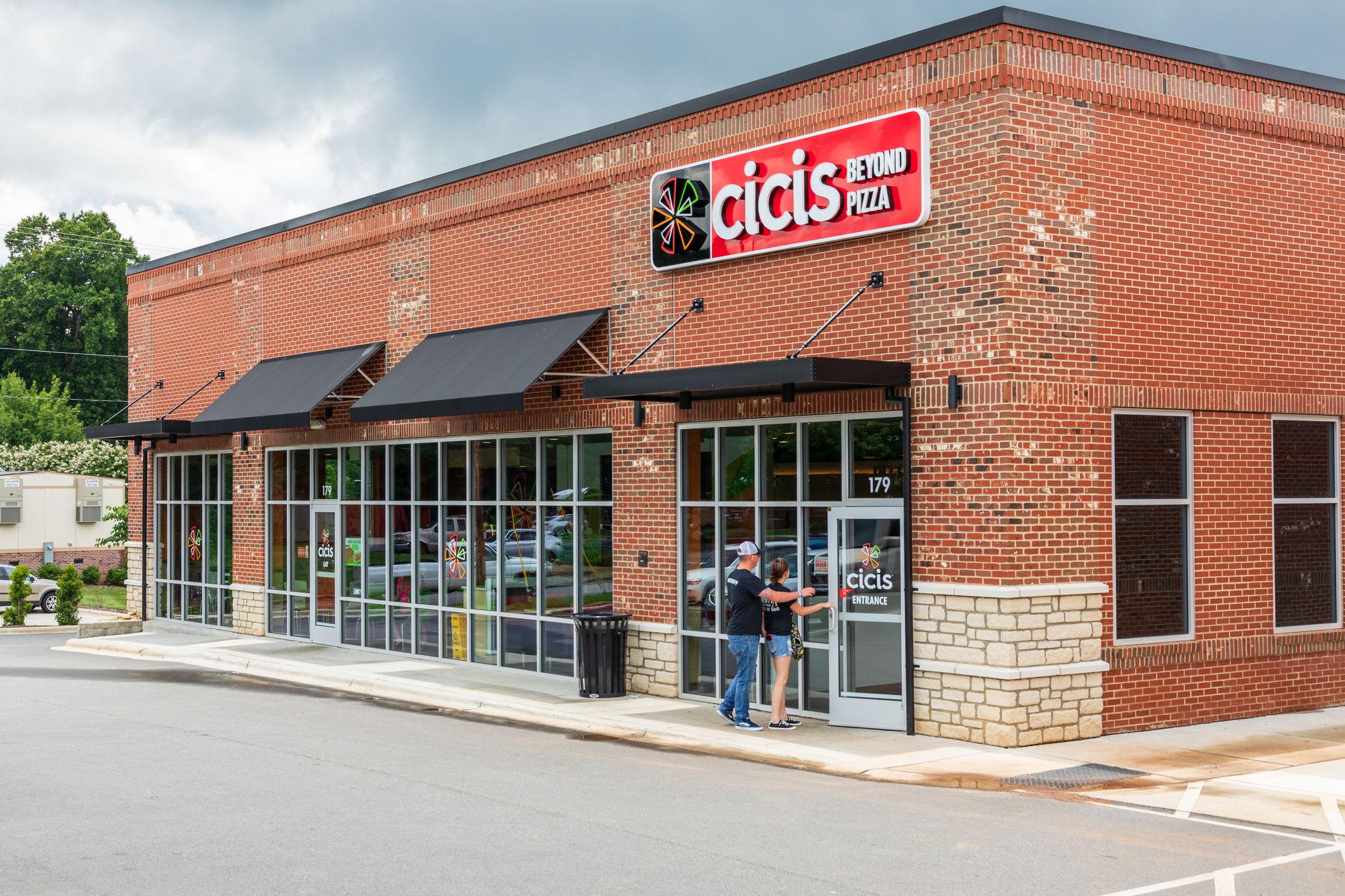 Front and side exterior of Cicis, Beyond Pizza Restaurant, Statesville, North Carolina, brick building, two people opening the front door going inside, driveway in the foreground, on a sunny day with grey clouds
