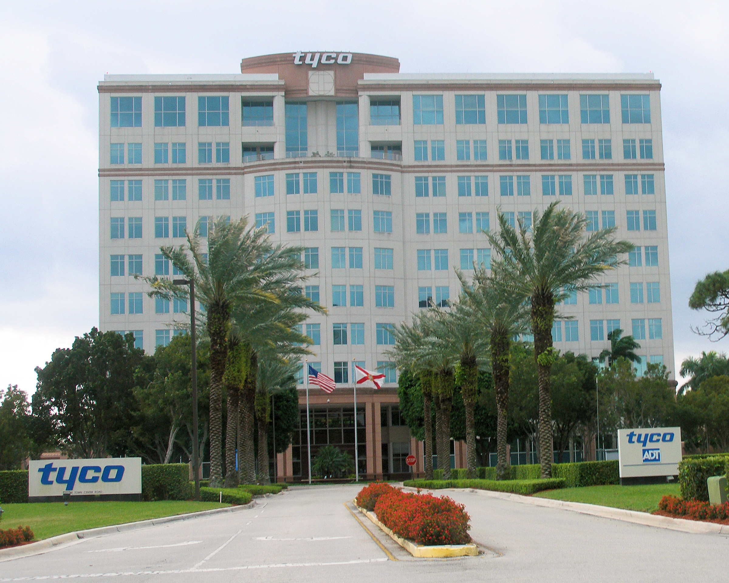 Front exterior of Tyco Fire & Security headquarters in Boca Raton, Florida, drive-thru road in the foreground, large building in the background framed by palm and other trees, against a light blue clouded sky