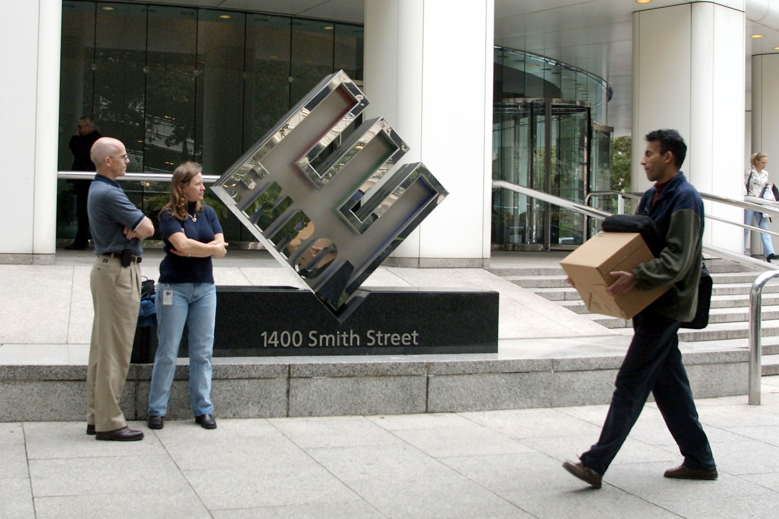Front of Enron headquarters, Houston, an employee on the right holds a box, is being laid off, two employees stand in front of the logo on the left, people are walking up and down the stairs in the background on the right