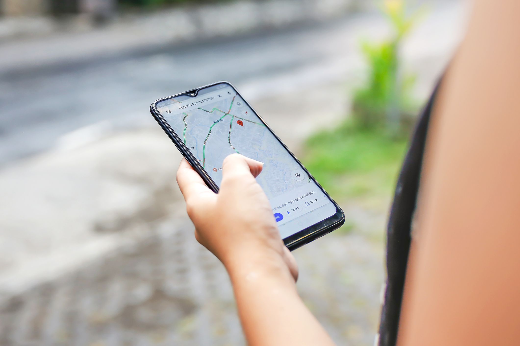 Hand holding an android smartphone, selective focus, map app opened on smartphone, person on the right, sidewalk, greenery, and street blurred in the background