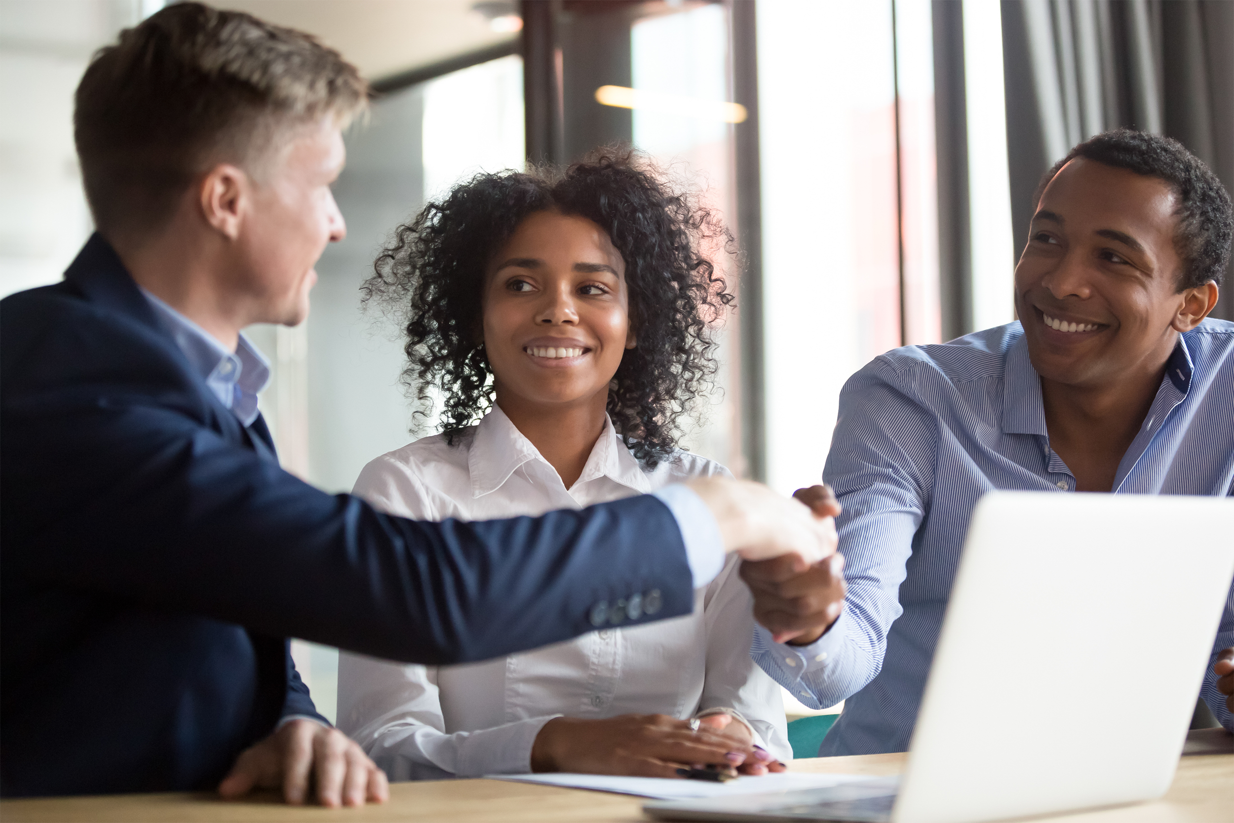 Man on the left shaking hand of man of couple on the right, woman is in the middle, all are smiling and sitting at a table with a laptop, couple is looking at man on the left, windows in an office blurred in the background