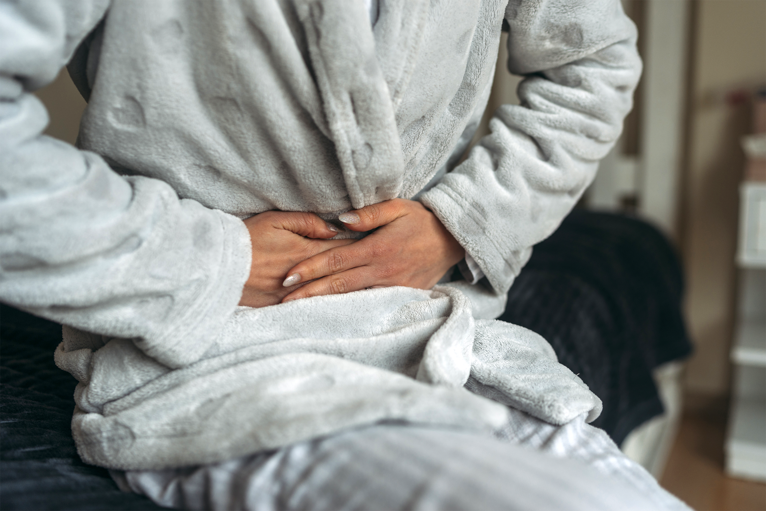 Mid-section of woman in distress holding her abdominal, wearing a light grey velour robe and long light grey pajama pants, while sitting on bed, blurred, bedroom is blurred in the background